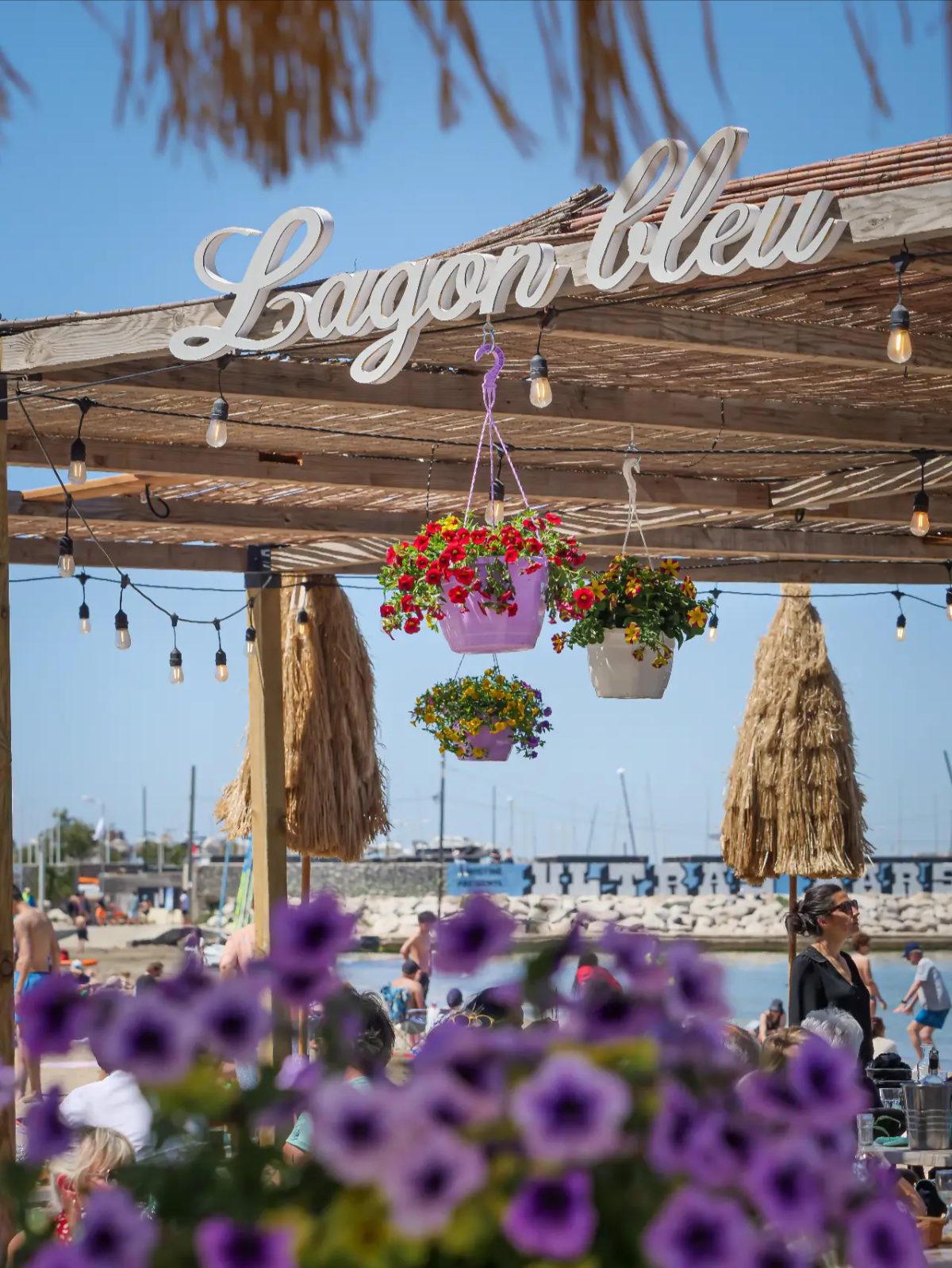Enseigne « Lagon bleu » avec parasols paille et fleurs roses face à la mer